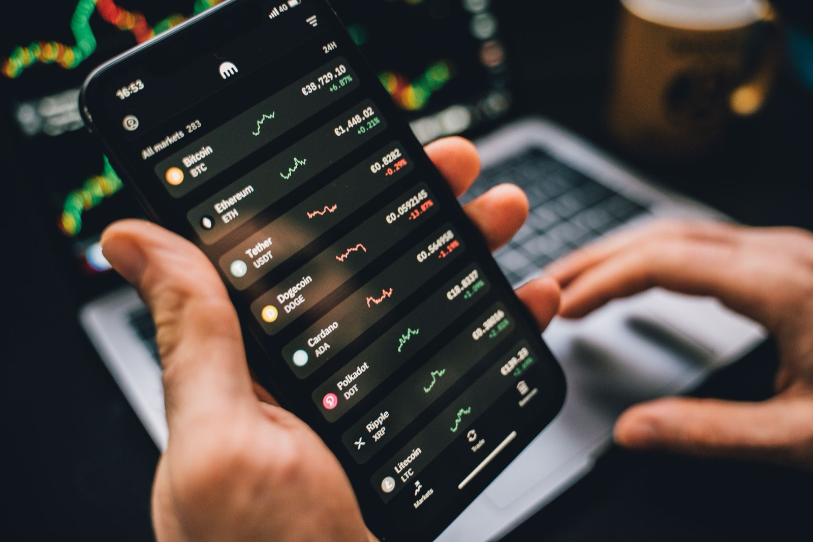 A hand holding a smartphone showing cryptocurrency prices above a laptop